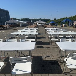 PERSEID SPÓŁKA Z OGRANICZONĄ ODPOWIEDZIALNOŚCIĄ - Ustawione rzędy białych stołów i krzeseł na zewnątrz, w tle namioty i budynek, sugerujące przygotowania do imprezy plenerowej lub targów.