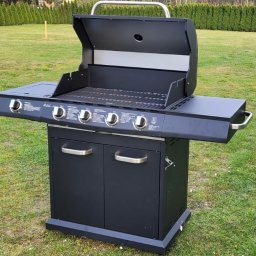 PERSEID SPÓŁKA Z OGRANICZONĄ ODPOWIEDZIALNOŚCIĄ - Czarny grill gazowy z czterema palnikami, otwartą pokrywą i szafką dolną na trawniku. Widoczne pokrętła regulacji i boczne półki. Grill gotowy do użycia na imprezie plenerowej.