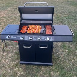 PERSEID SPÓŁKA Z OGRANICZONĄ ODPOWIEDZIALNOŚCIĄ - Czarny grill gazowy na trawie z przygotowanymi szaszłykami, stekami i kiełbasami. Grill otwarty, gotowy do użycia. Akcesoria do grilla obok.