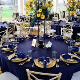 Uroczysty stół z granatowym obrusem, złotymi talerzami i bukietami żółtych kwiatów. Eleganckie krzesła i dekoracje podkreślają wyjątkowy charakter wydarzenia.
