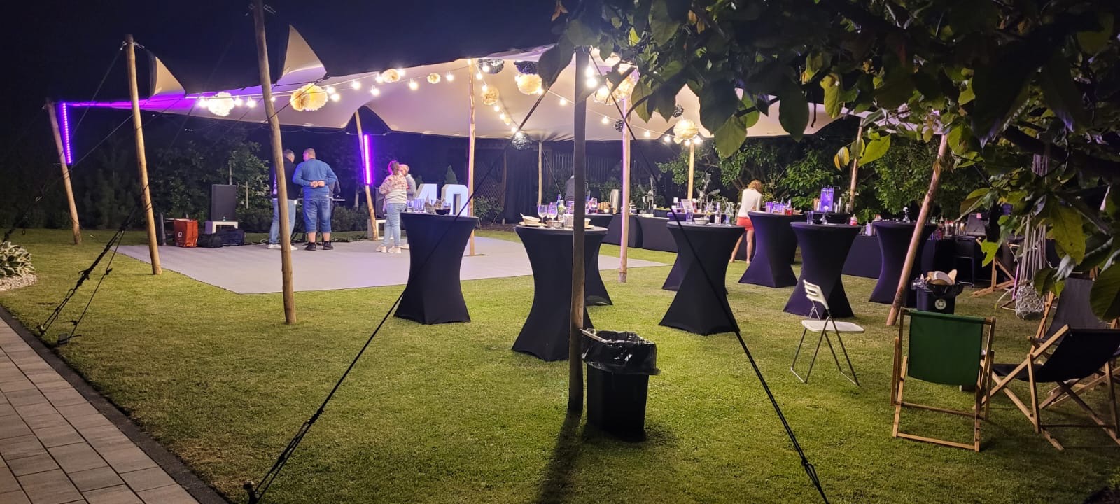 Nocna impreza plenerowa pod białym namiotem afrykańskim z oświetleniem girlandami żarówek, czarne stoliki koktajlowe, parkiet taneczny i dekoracje świetlne LED w ogrodzie.
