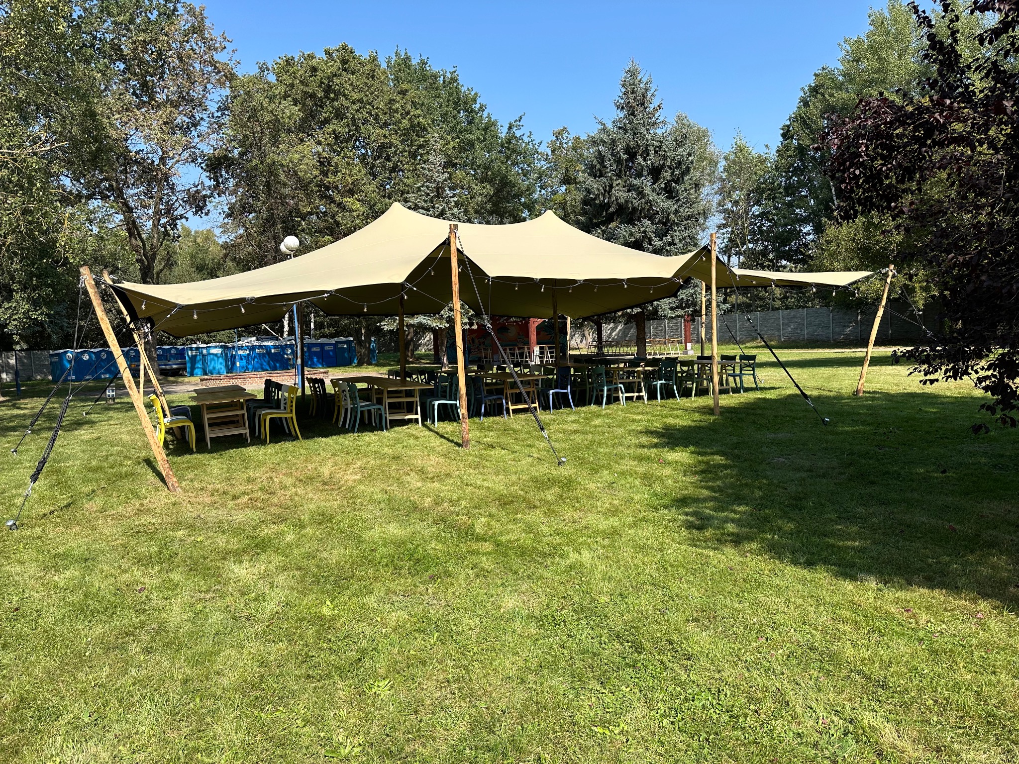 Rozłożysty, beżowy namiot plenerowy z drewnianymi słupami, oświetlony girlandami żarówek, ustawiony na trawniku, z widocznymi stołami i krzesłami wewnątrz, oraz przenośnymi toaletami w tle.