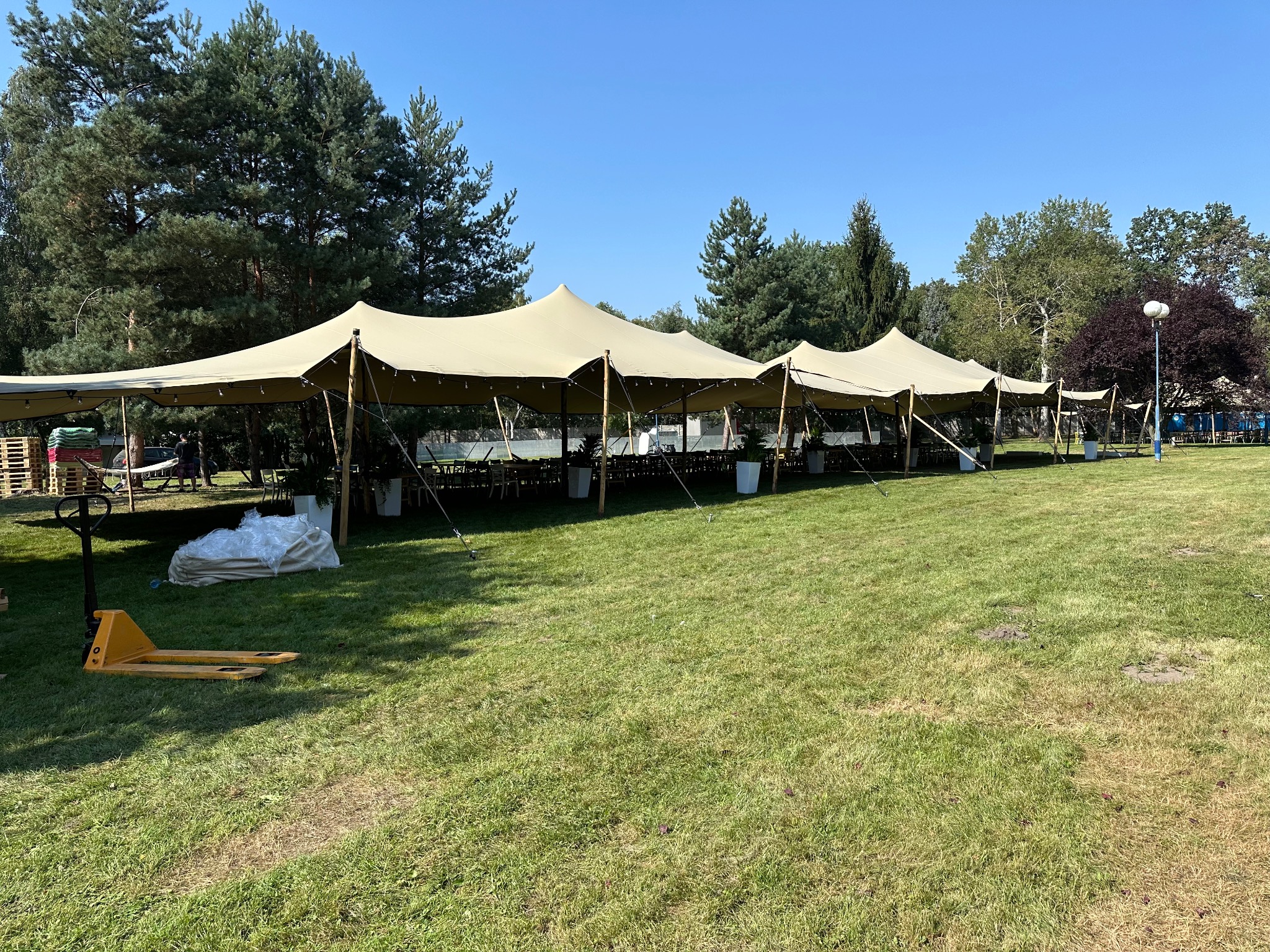Rozstawione rzędy beżowych namiotów plenerowych z oświetleniem, stołami i krzesłami, na zielonym trawniku, z paletami i wózkiem paletowym w pierwszym planie, w tle drzewa i błękitne niebo.