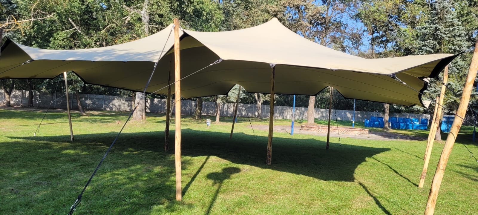 Beżowy namiot plenerowy rozstawiony na trawniku, drewniane słupy nośne, widoczne linie naciągowe i fragment murku w tle.