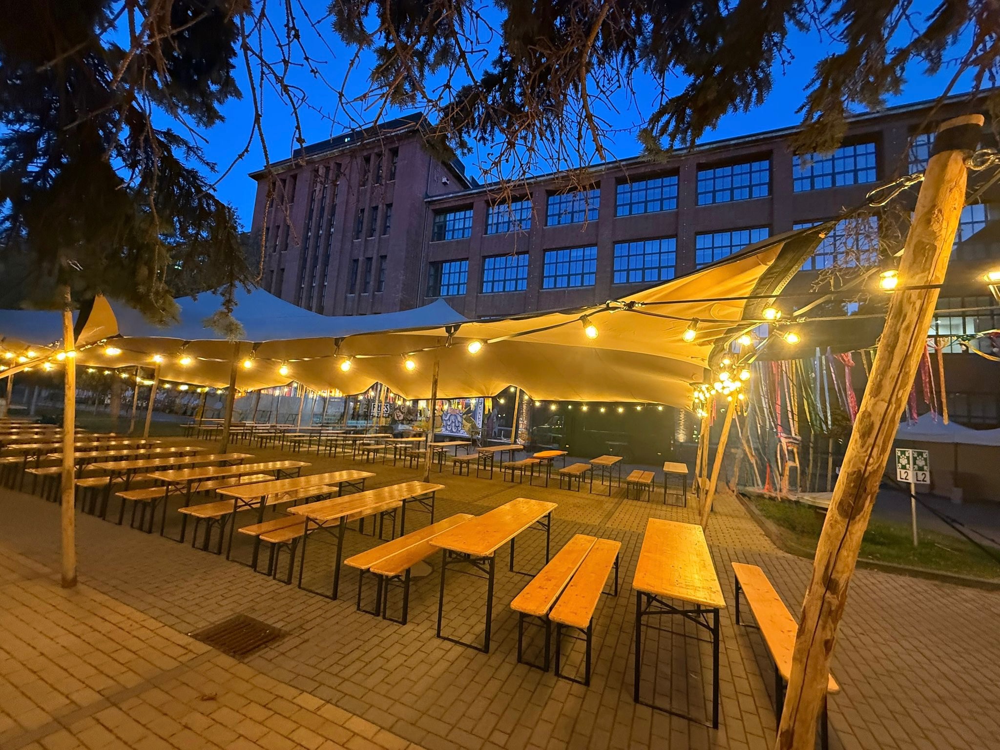 Oświetlony namiot imprezowy z drewnianymi stołami i ławkami, ustawiony na zewnątrz budynku. Dekoracyjne girlandy żarówkowe tworzą ciepłą atmosferę.