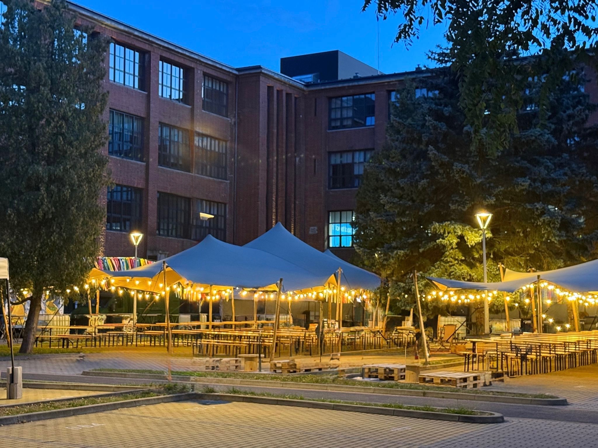 Oświetlony teren eventowy z namiotami i drewnianymi stołami na zewnątrz budynku z cegły, wieczorową porą. Dekoracyjne girlandy świetlne, palety jako podesty.