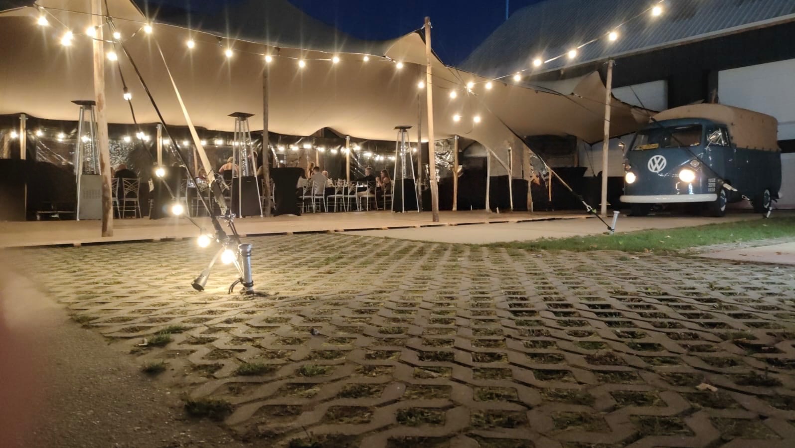 Oświetlony namiot eventowy z dekoracyjnymi żarówkami, grzejnikami i zaparkowanym Volkswagenem Transporterem w tle, wieczorna sceneria.
