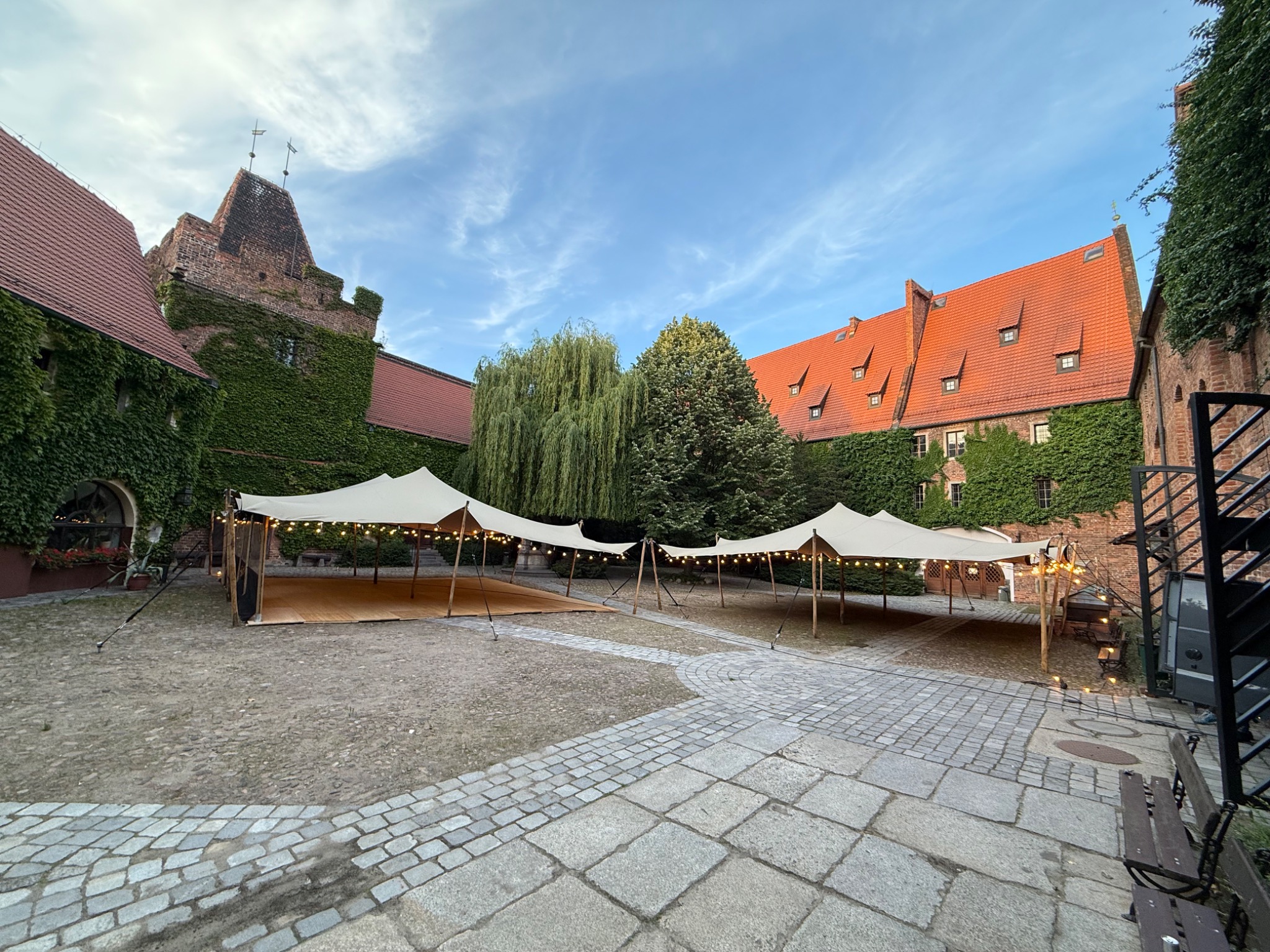 Dwa rozstawione namioty z oświetleniem na dziedzińcu zamku obrośniętego bluszczem, przygotowane na imprezę plenerową. Kamienna nawierzchnia i dekoracyjne detale architektoniczne.