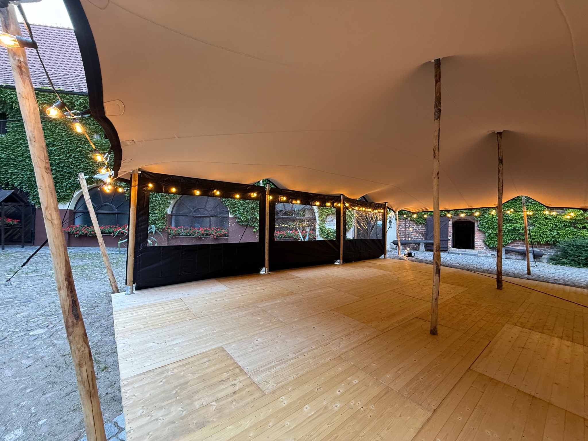 Biały namiot plenerowy ze ścianami, drewnianą podłogą i girlandami żarówek, ustawiony na kamiennym dziedzińcu z budynkiem porośniętym bluszczem w tle.