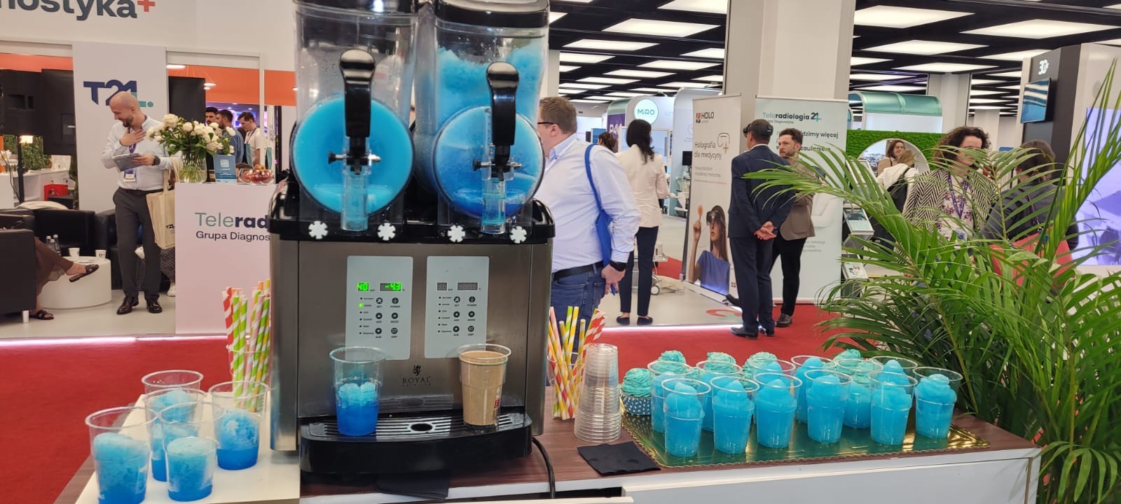 Automat do mrożonych napojów na stoisku targowym z niebieskimi slushami w kubkach. W tle widoczni ludzie i stoiska.