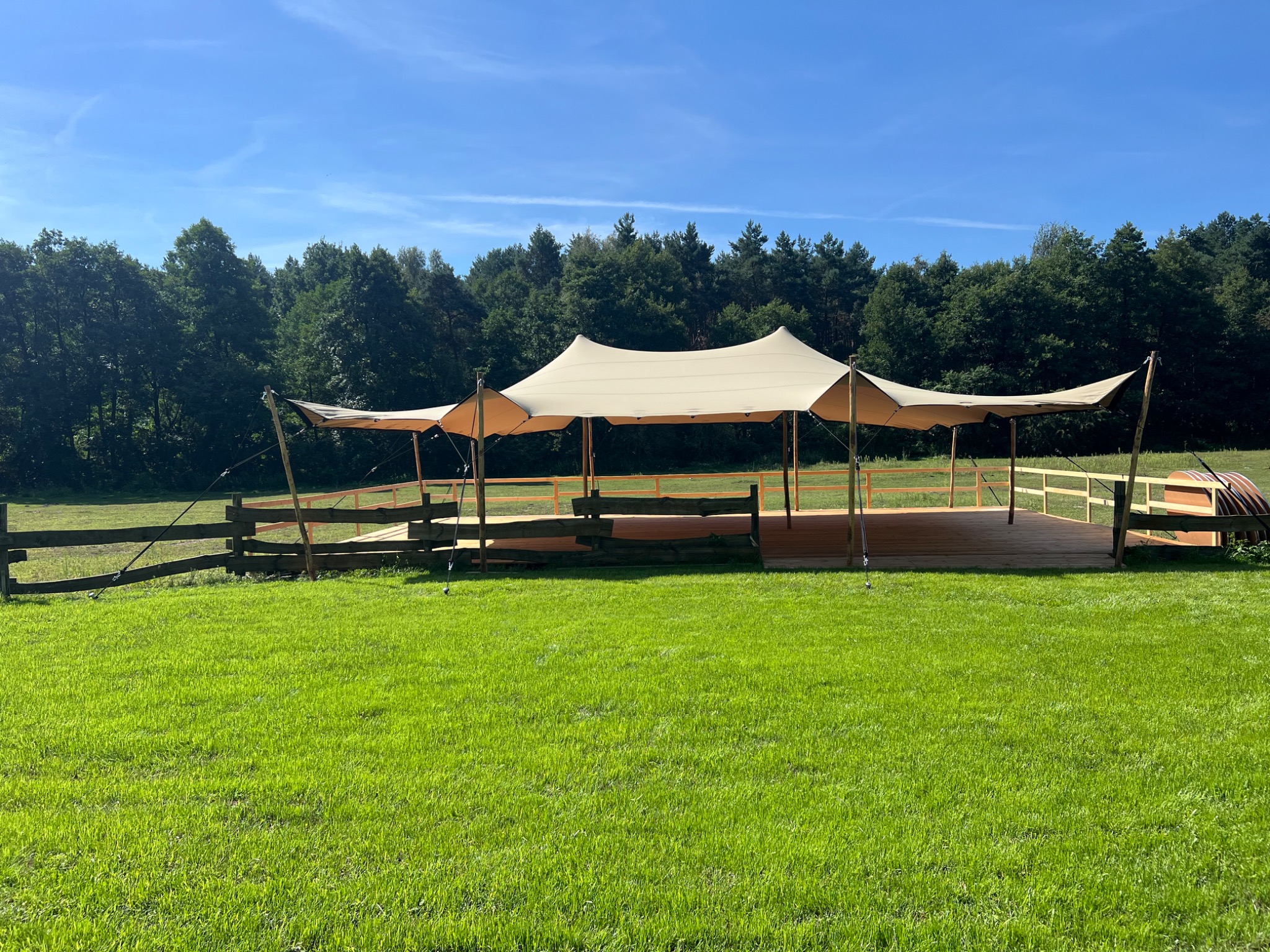 Rozłożysty namiot plenerowy z drewnianym podestem na zielonej łące, otoczony lasem pod błękitnym niebem. Widok z daleka, prezentujący konstrukcję i otoczenie.