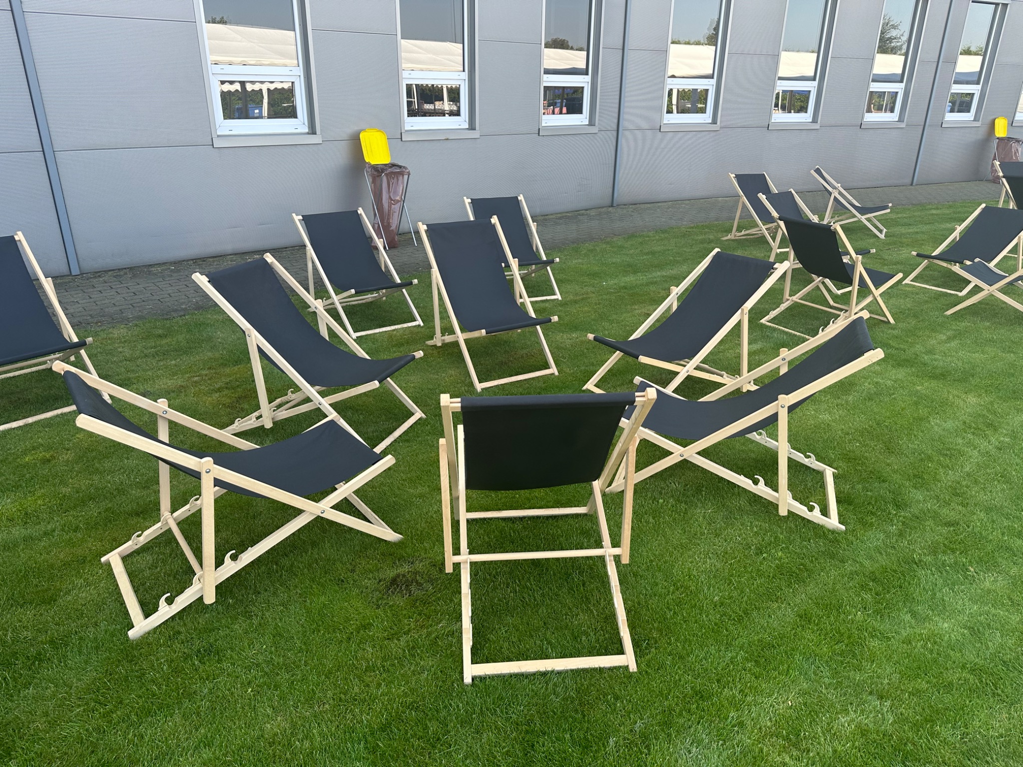 Rozstawione leżaki plażowe z czarnym materiałem na trawie przed budynkiem z oknami. Konfiguracja gotowa na wydarzenie plenerowe.