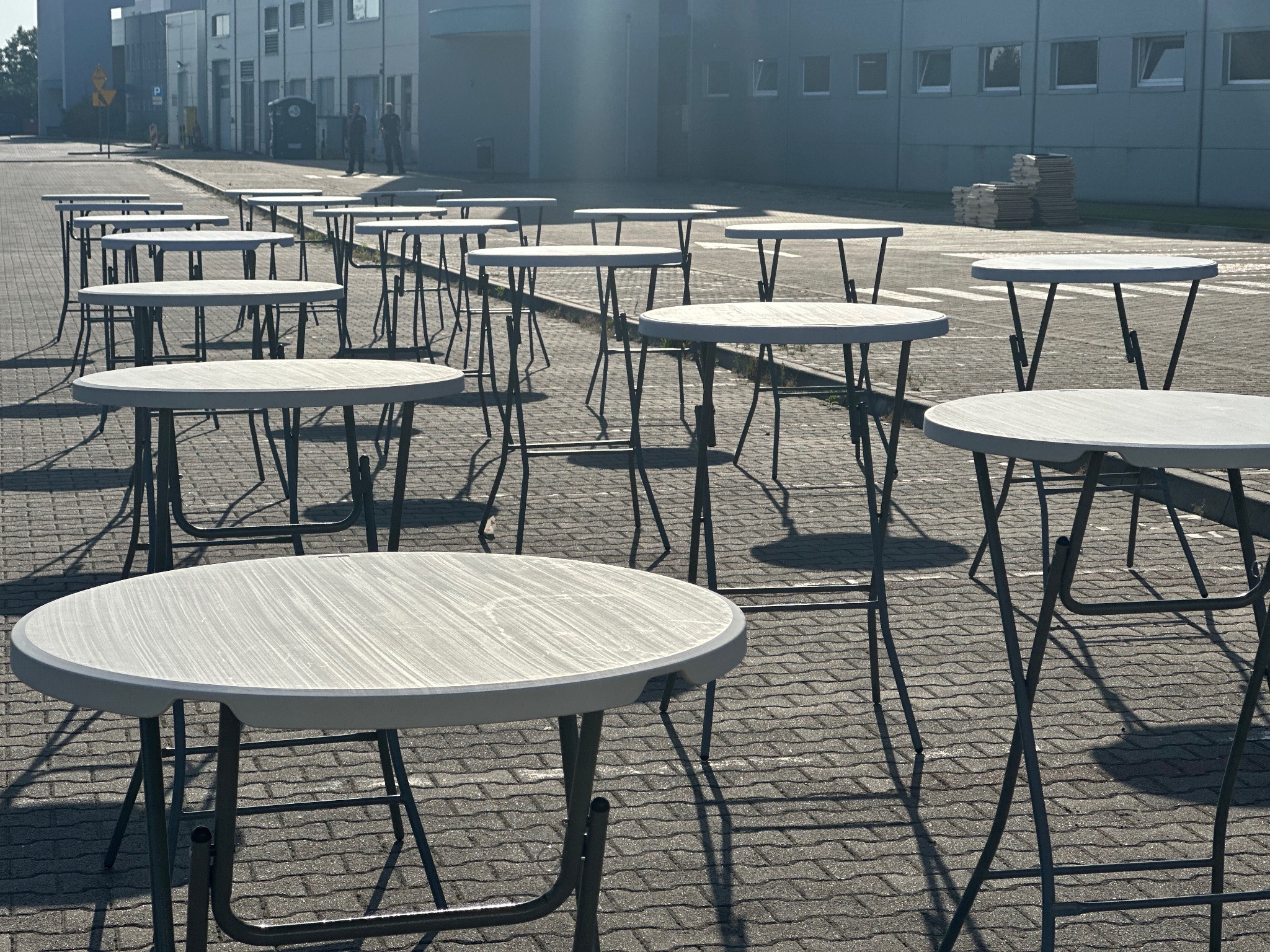 Rząd białych, okrągłych stolików koktajlowych z metalowymi nogami, ustawionych na zewnątrz na brukowanym placu przed budynkiem. Cienie i ostre światło słoneczne.