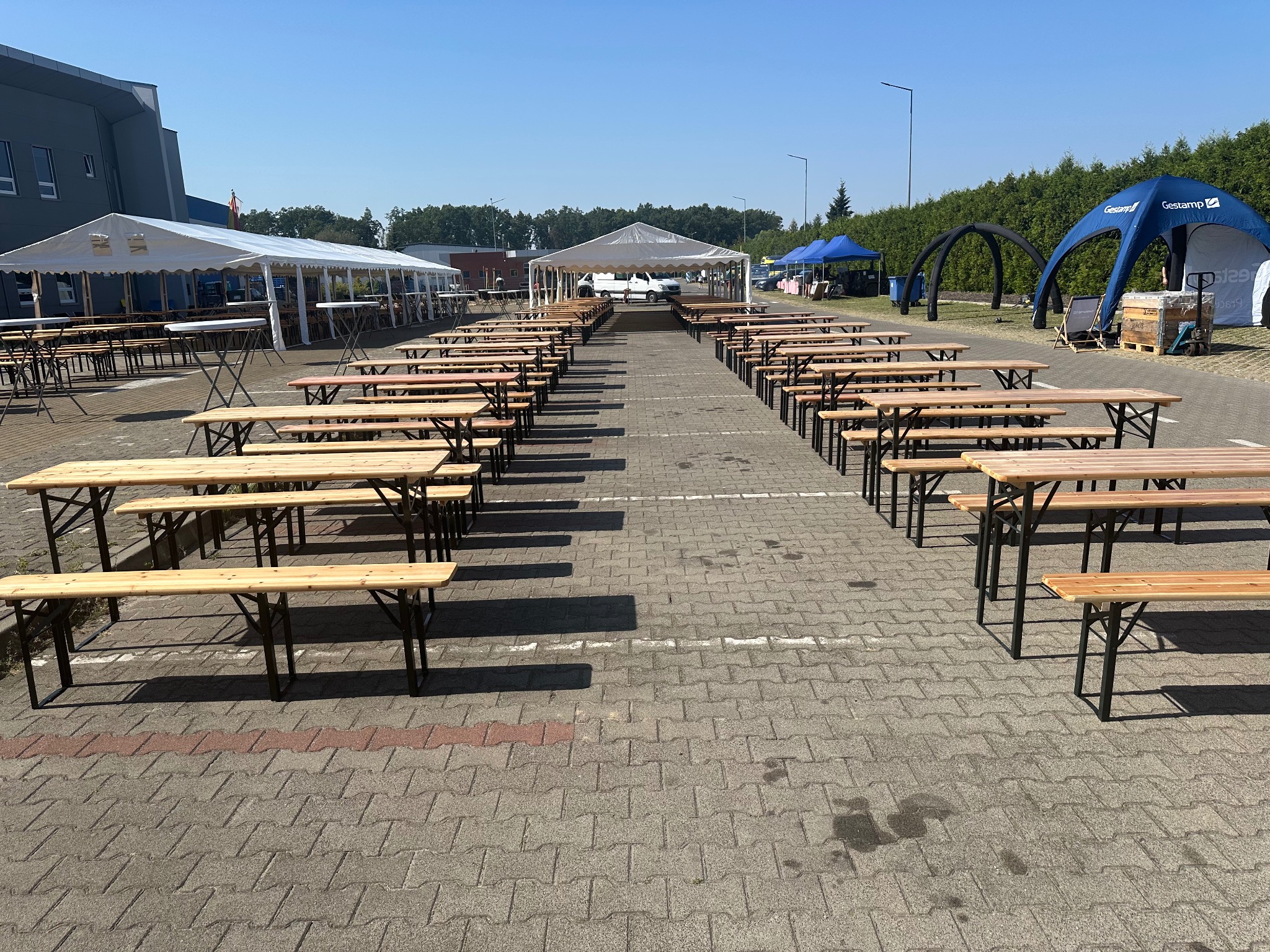 Ustawione rzędy drewnianych stołów i ławek, przygotowane do imprezy plenerowej. W tle białe i niebieskie namioty eventowe.