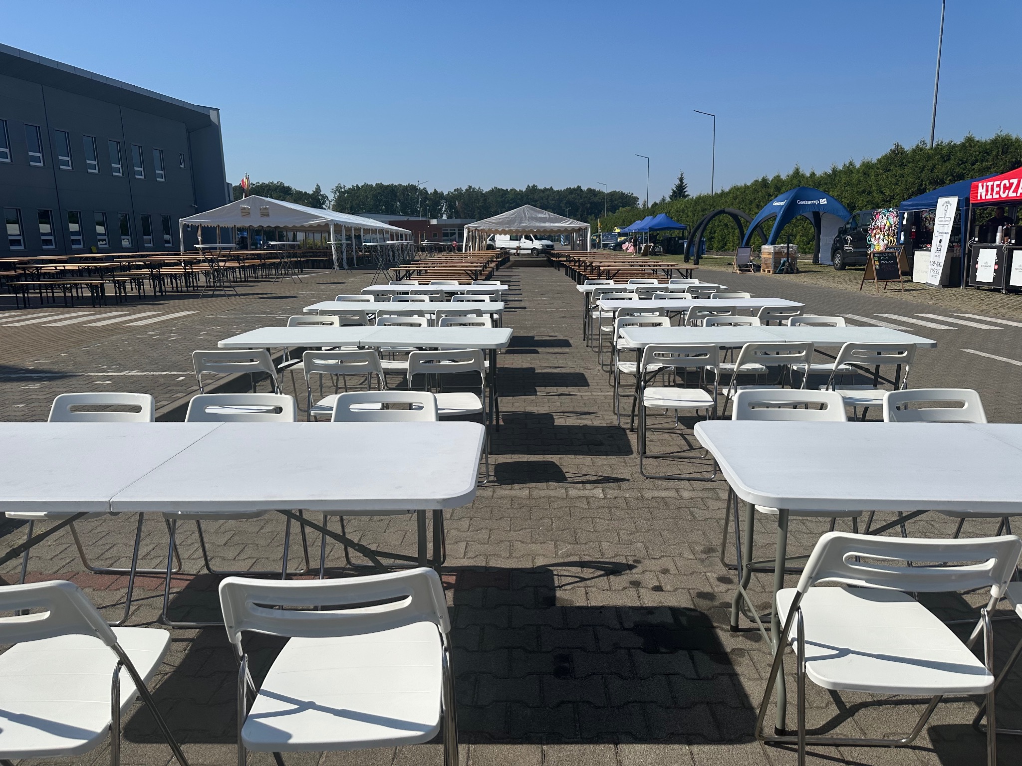 Ustawione rzędy białych stołów i krzeseł na zewnątrz, w tle namioty i budynek, sugerujące przygotowania do imprezy plenerowej lub targów.