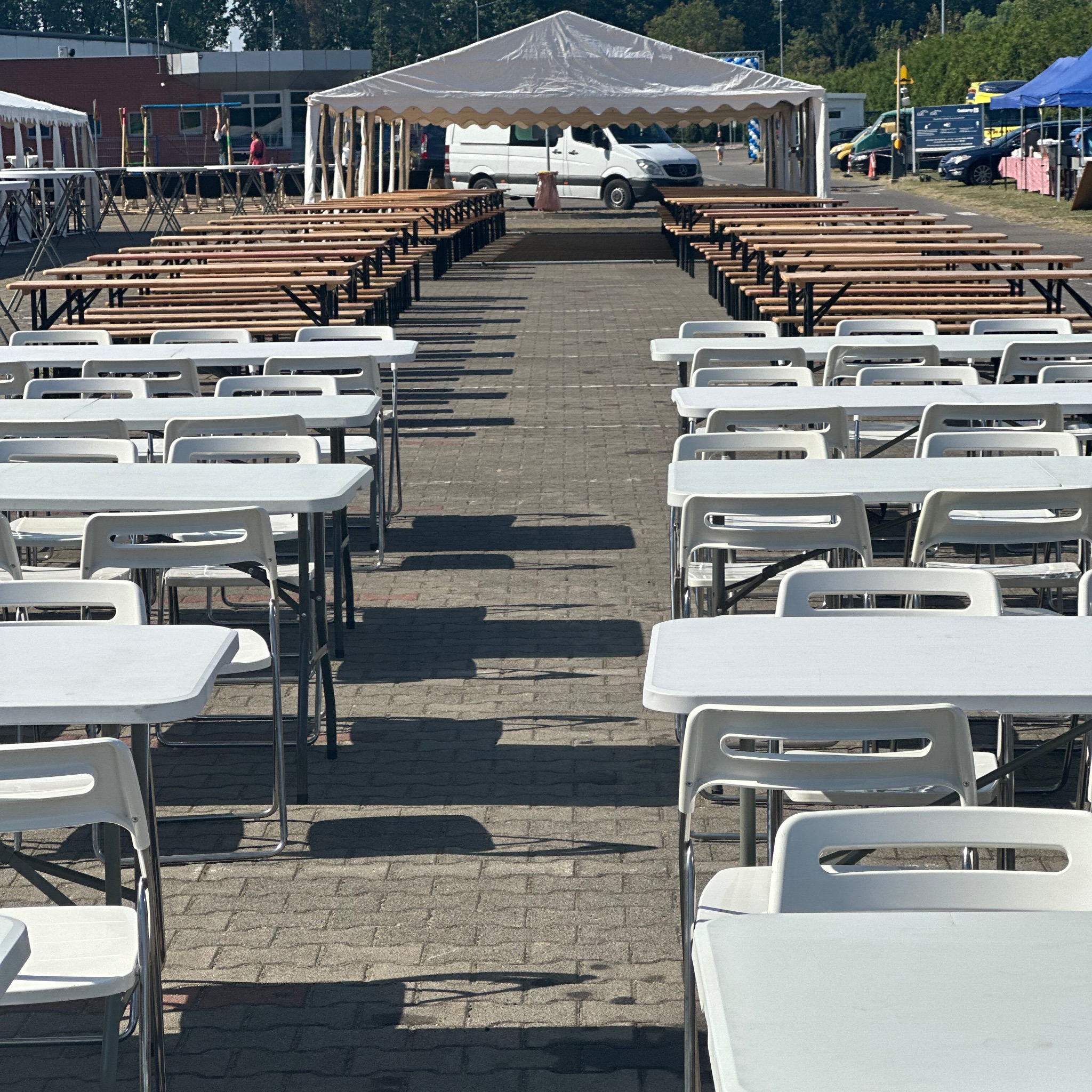 Ustawione rzędy białych stołów i krzeseł prowadzące do namiotu z białym vanem w środku, sugerujące przygotowania do imprezy plenerowej.