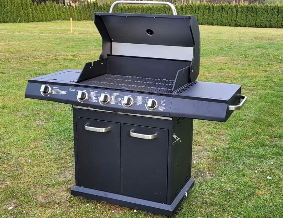 Czarny grill gazowy z czterema palnikami, otwartą pokrywą i szafką dolną na trawniku. Widoczne pokrętła regulacji i boczne półki. Grill gotowy do użycia na imprezie plenerowej.