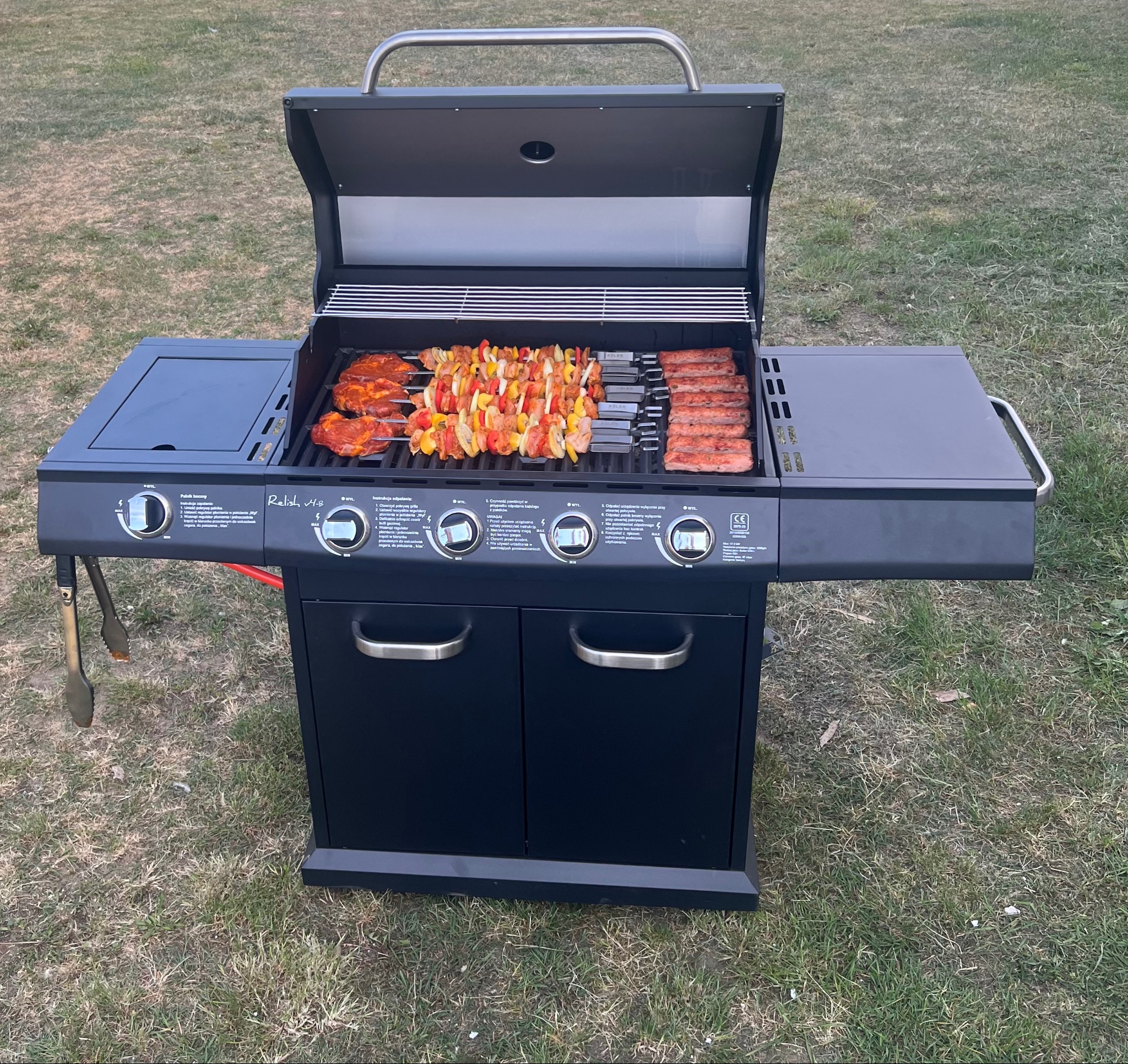 Czarny grill gazowy na trawie z przygotowanymi szaszłykami, stekami i kiełbasami. Grill otwarty, gotowy do użycia. Akcesoria do grilla obok.