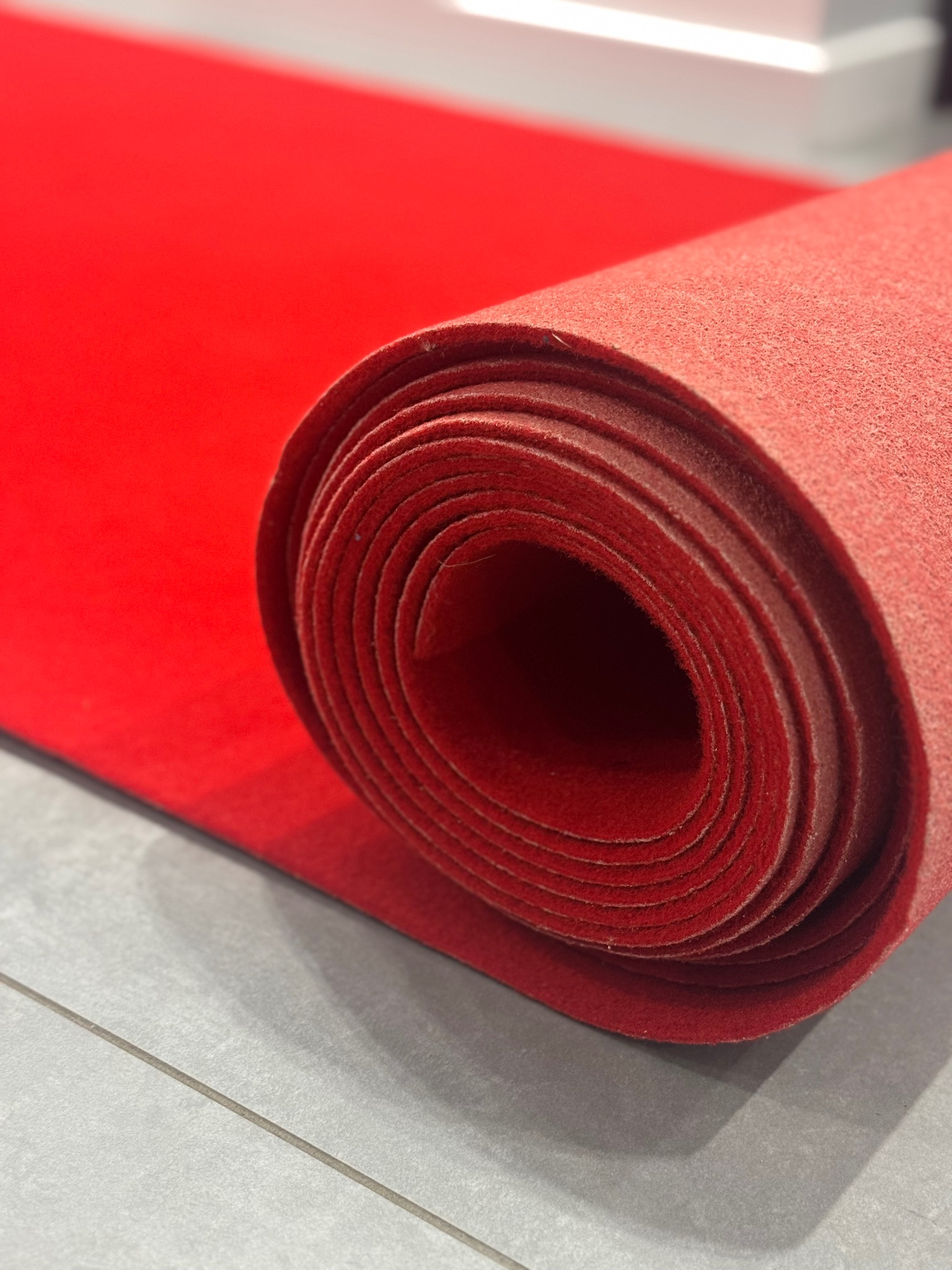 Czerwony, zwinięty dywan na szarych płytkach podłogowych, oświetlony górnym światłem, sugerujący przygotowanie do eventu lub wynajem dywanów.