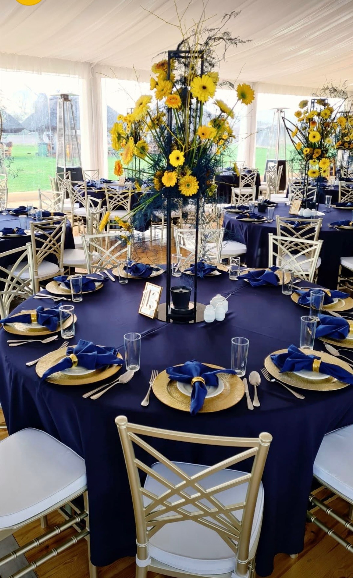 Uroczysty stół z granatowym obrusem, złotymi talerzami i bukietami żółtych kwiatów. Eleganckie krzesła i dekoracje podkreślają wyjątkowy charakter wydarzenia.
