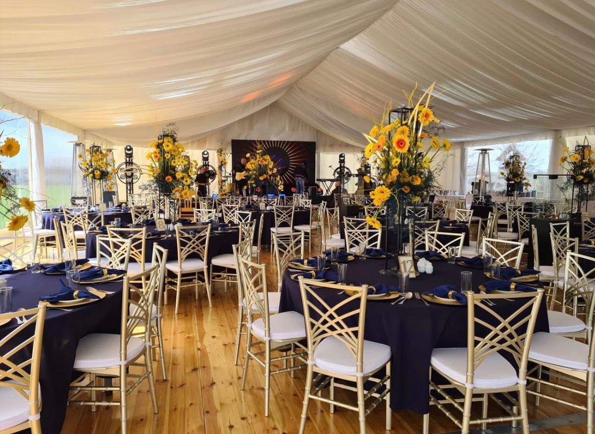 Uroczysta aranżacja wnętrza namiotu eventowego: okrągłe stoły z ciemnymi obrusami, złote krzesła, kwiatowe dekoracje i oświetlenie sceniczne w tle.