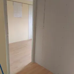 Montaż płyt kartonowo-gipsowych na ścianie, widoczne łączenia płyt, w tle fragment pokoju z panelami podłogowymi i zasłonami.