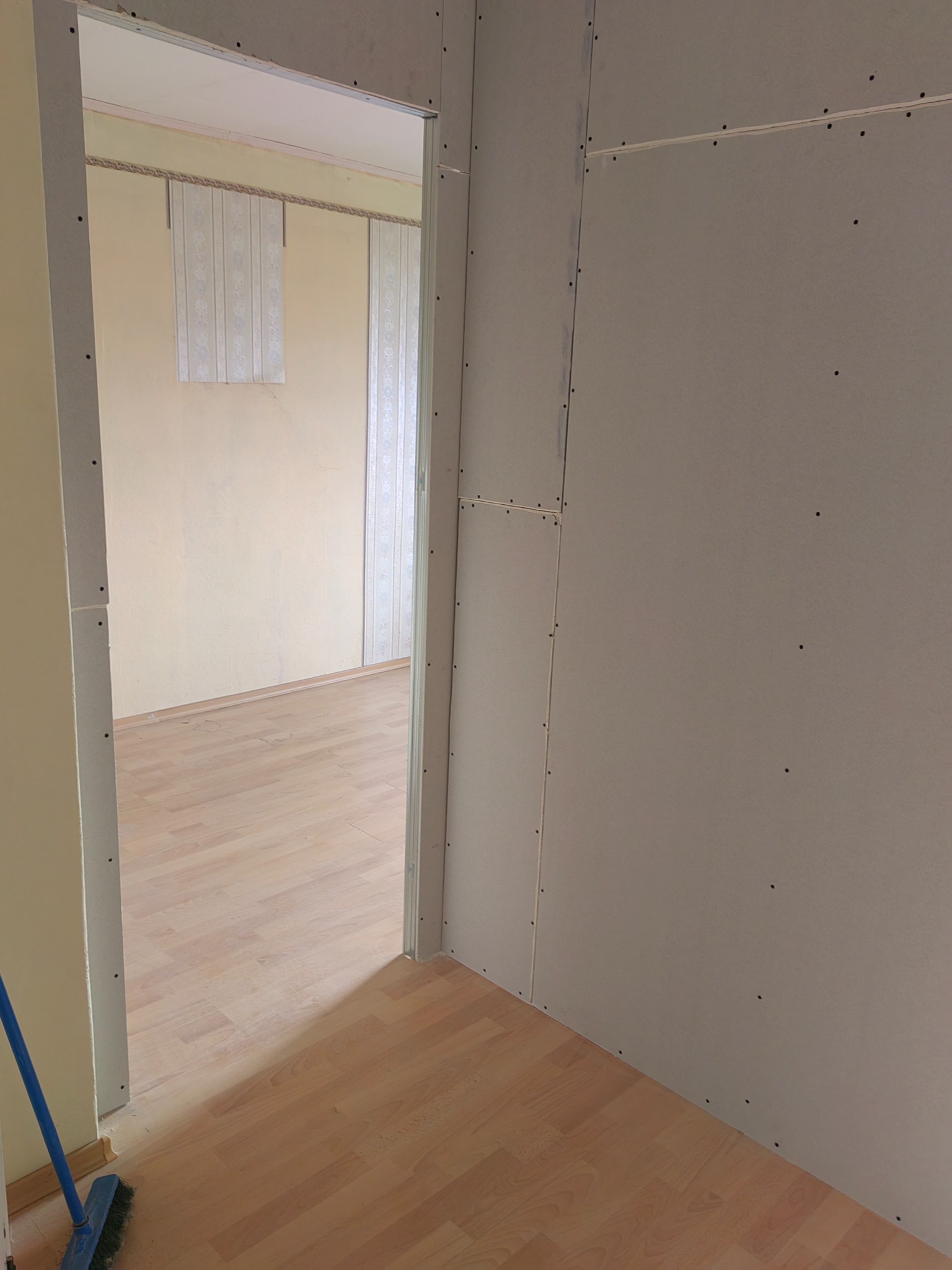 Montaż płyt kartonowo-gipsowych na ścianie, widoczne łączenia płyt, w tle fragment pokoju z panelami podłogowymi i zasłonami.