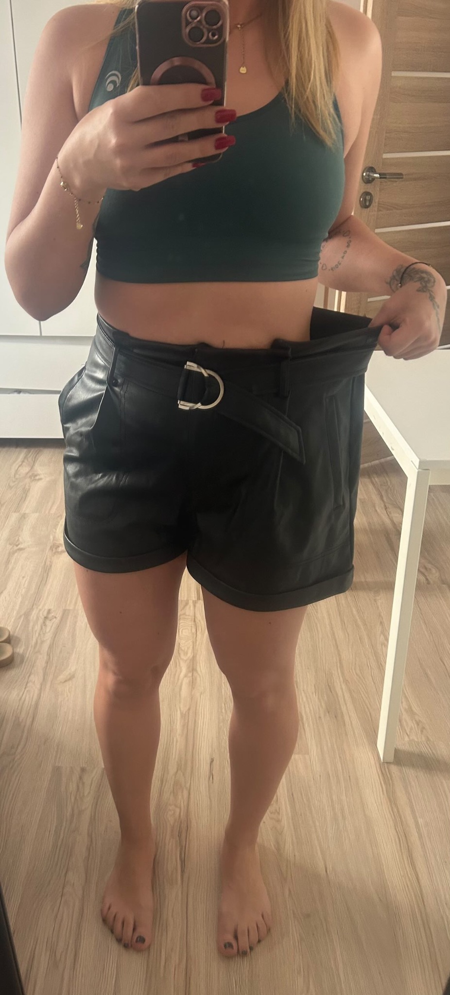 Kobieta w sportowym topie i skórzanych szortach, trzymająca luźny pas, demonstrująca utratę wagi, widoczny tatuaż na ramieniu.