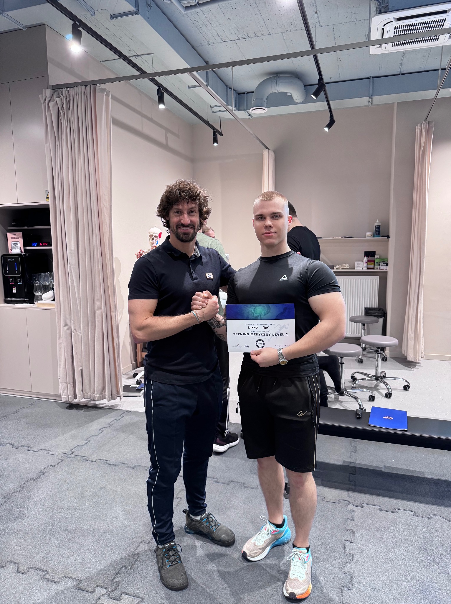 Uśmiechnięty mężczyzna w sportowym stroju ściska dłoń drugiemu mężczyźnie, który trzyma certyfikat ukończenia 'Treningu Medycznego Level 2', w tle widoczne wyposażenie siłowni.