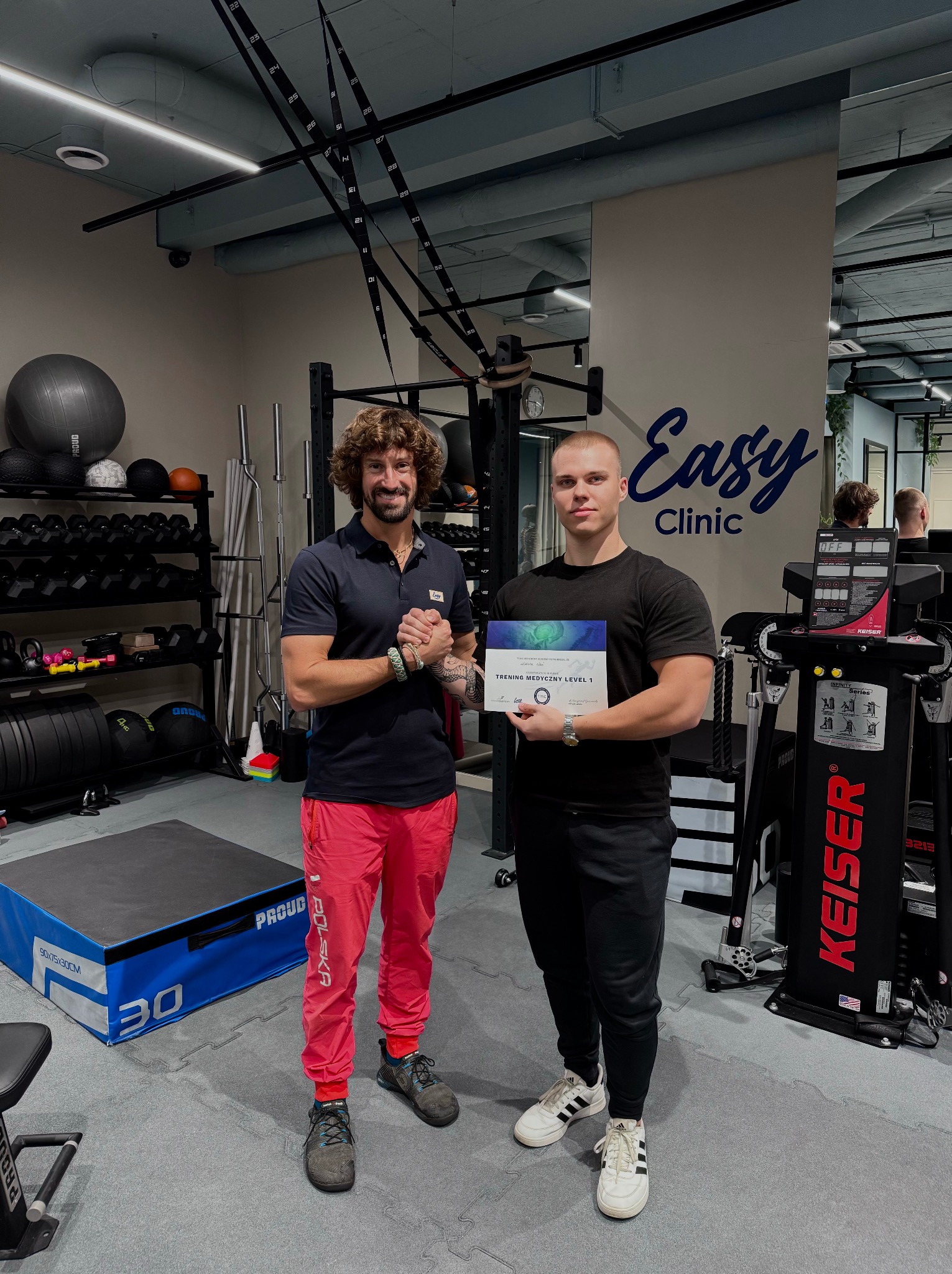 Dwóch mężczyzn w siłowni, jeden w czerwonych spodniach i z brodą, drugi trzyma certyfikat 'Trening Medyczny Level 1' przed maszyną Keiser i logo 'Easy Clinic'.