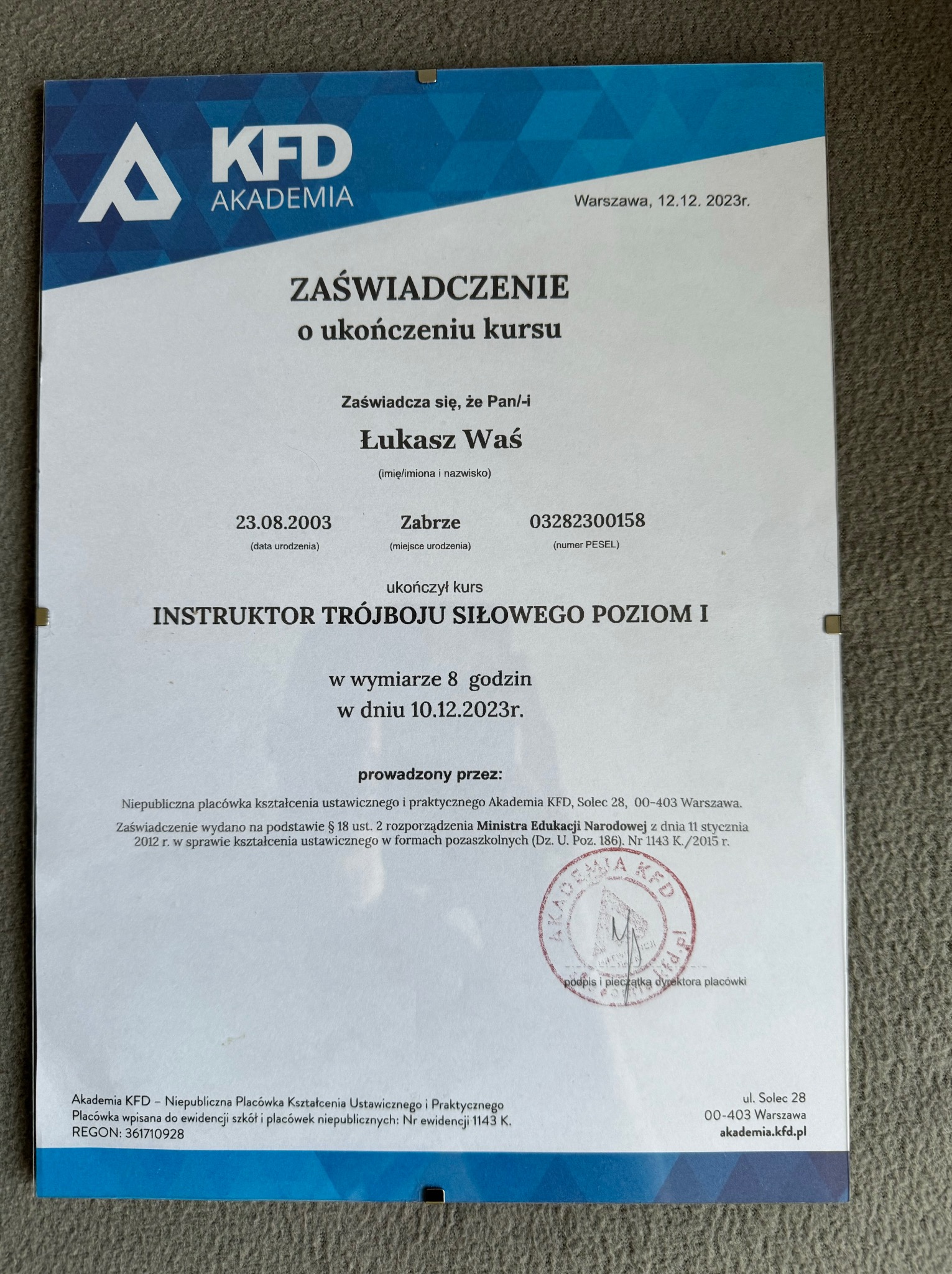 Skan zaświadczenia o ukończeniu kursu instruktora trójboju siłowego poziomu I, wydanego przez Akademię KFD, z datą 12.12.2023, zawierające dane osobowe i pieczęć.