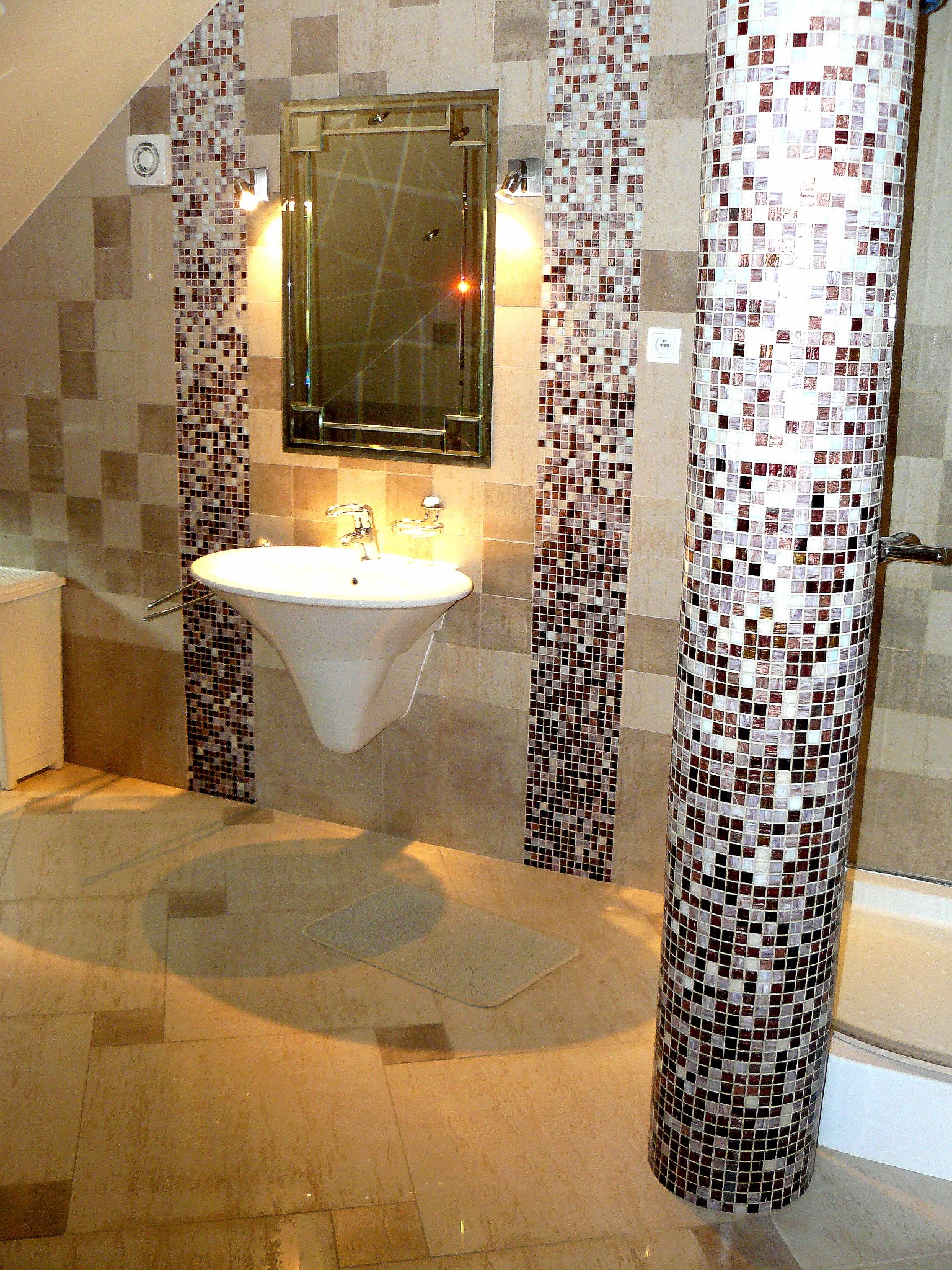 Umywalka wisząca z chromowaną armaturą i mozaiką w odcieniach brązu i beżu na ścianie i kolumnie w wykończonej łazience.