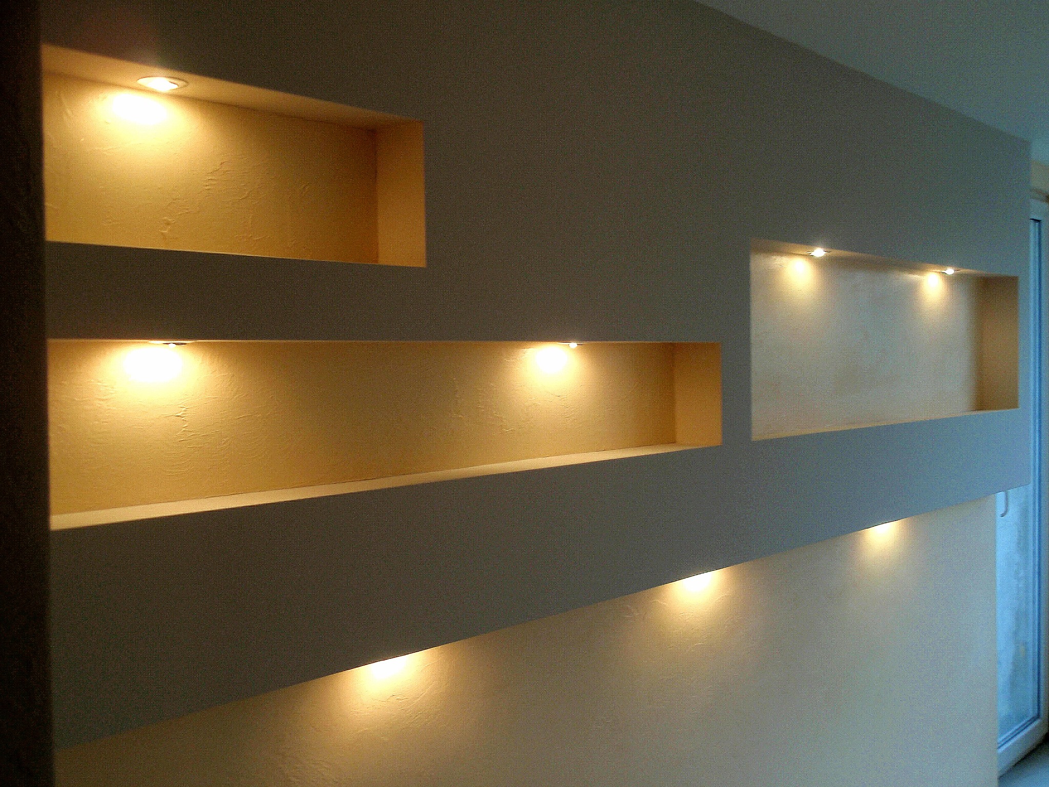 Ściana z wnękami podświetlonymi punktowym oświetleniem, faktura tynku dekoracyjnego w odcieniach beżu i szarości, nowoczesny design.