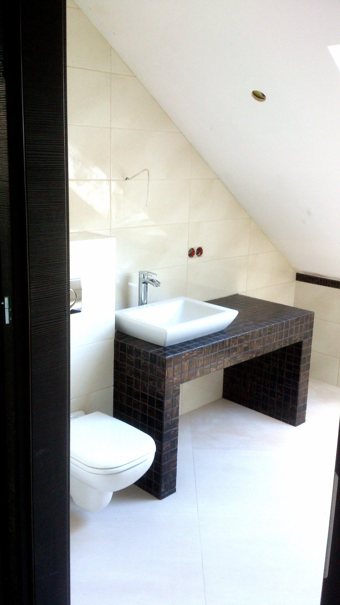 Ukośna łazienka na poddaszu z umywalką nablatową na postumencie z ciemnej mozaiki i podwieszaną toaletą, widok zza futryny drzwi.