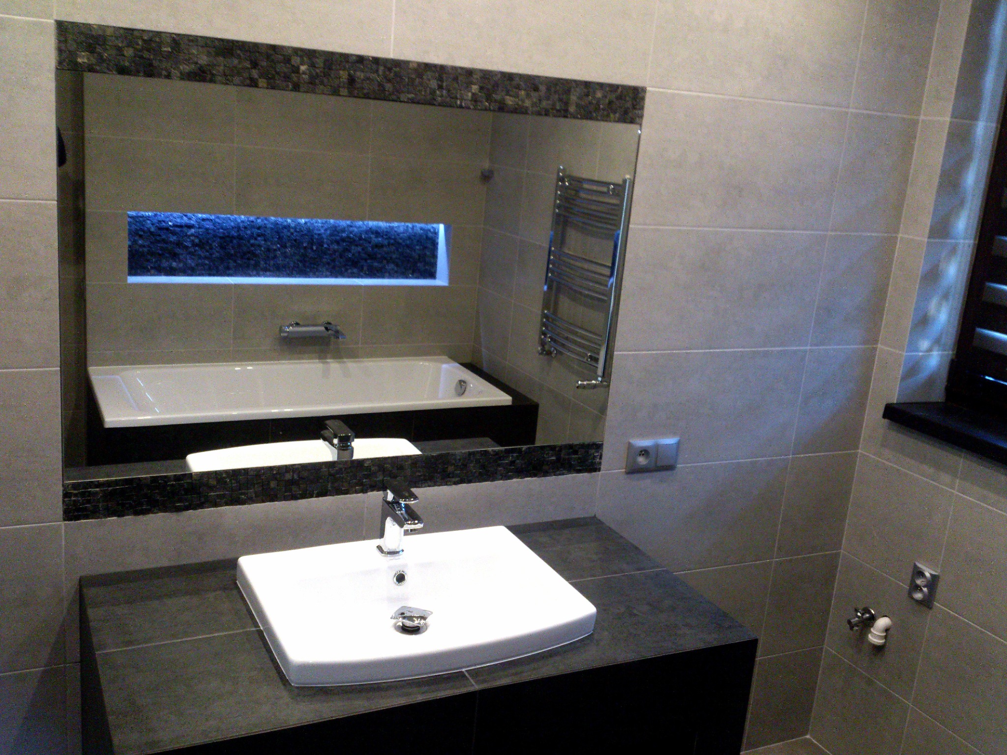 Umywalka nablatowa z chromowaną baterią na ciemnym blacie, w tle lustro z mozaikową ramą odbijające wannę i grzejnik drabinkowy w łazience wykończonej szarymi płytkami.