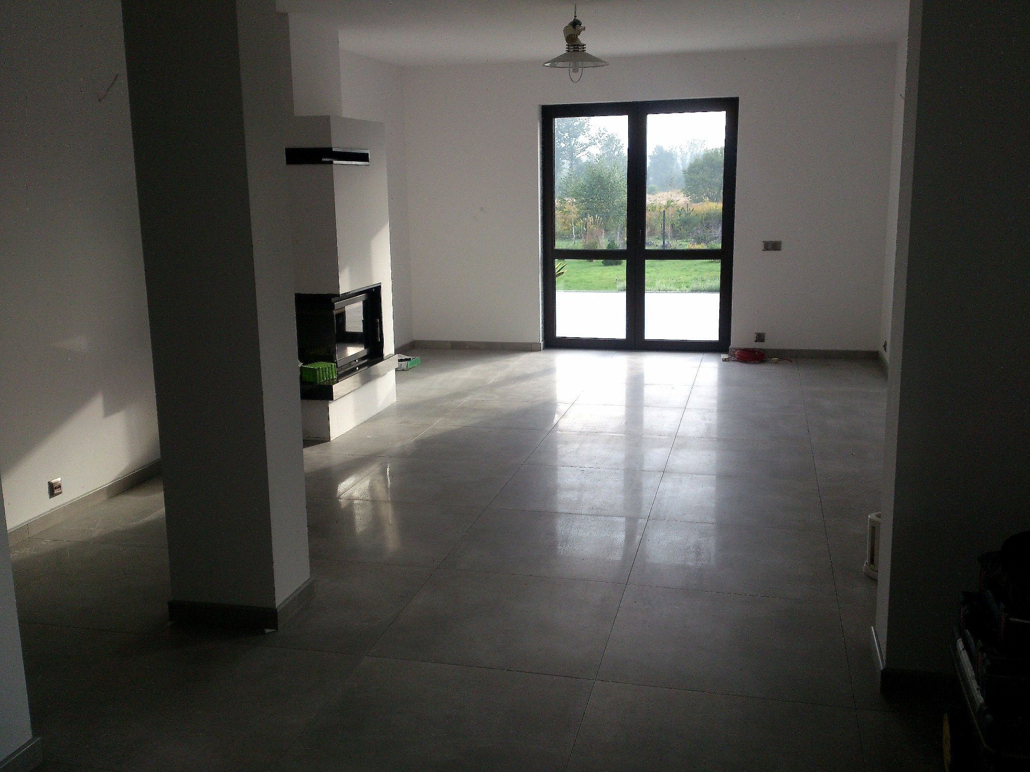 Przestronne, jasne wnętrze z szarą, błyszczącą posadzką z płytek, nowoczesnym kominkiem z czarnym wkładem, dużym oknem balkonowym z widokiem na zieleń i prostą lampą sufitową.