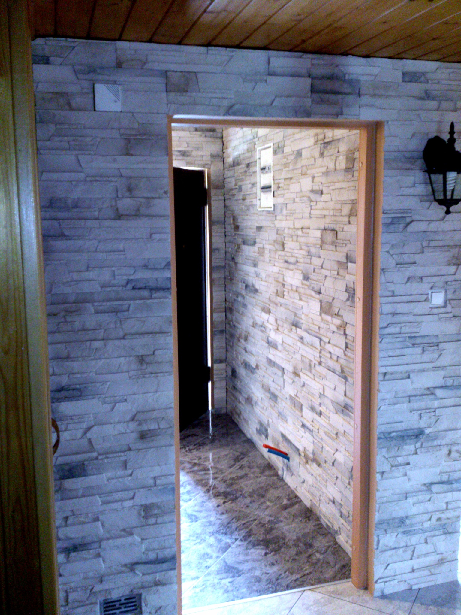 Ściana wyłożona dekoracyjną, szarą cegłą, widok na korytarz z marmurową podłogą i czarnymi drzwiami, drewniany sufit i ozdobny kinkiet.