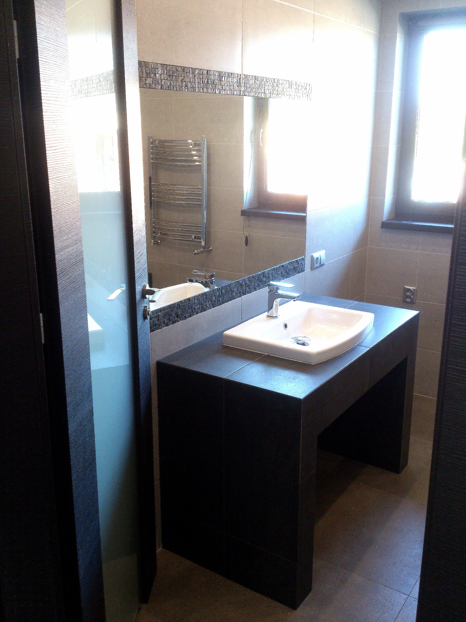 Nowoczesna łazienka z umywalką nablatową na czarnym, kafelkowym postumencie, widok na lustro z dekoracyjnym paskiem mozaiki, grzejnik drabinkowy i okno z naturalnym światłem.