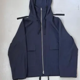 Niebieska kurtka z kapturem, zamek błyskawiczny na całej długości, dwie kieszenie z przodu, ściągacze u dołu i przy kapturze, leżąca na szarym tle.