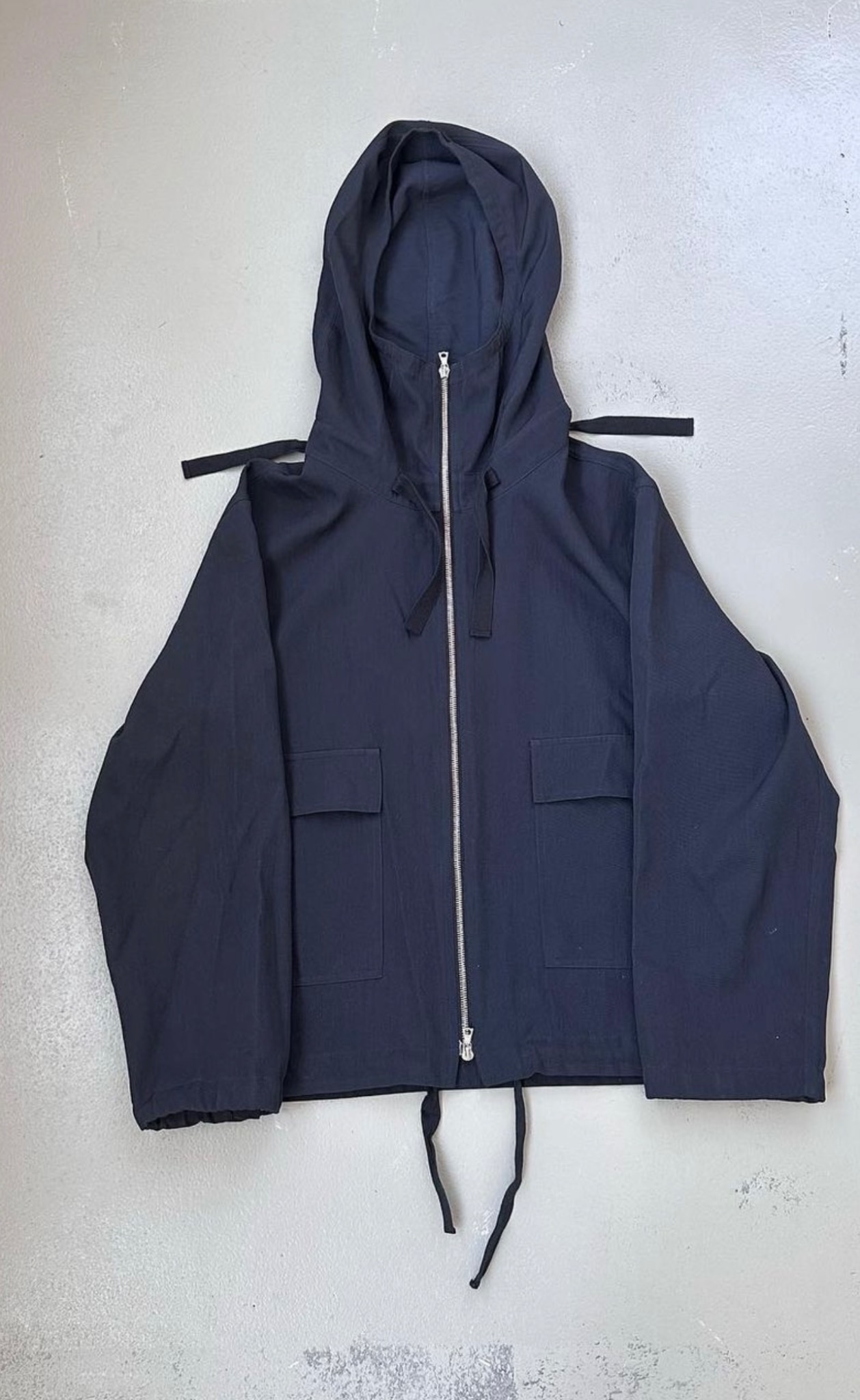 Niebieska kurtka z kapturem, zamek błyskawiczny na całej długości, dwie kieszenie z przodu, ściągacze u dołu i przy kapturze, leżąca na szarym tle.