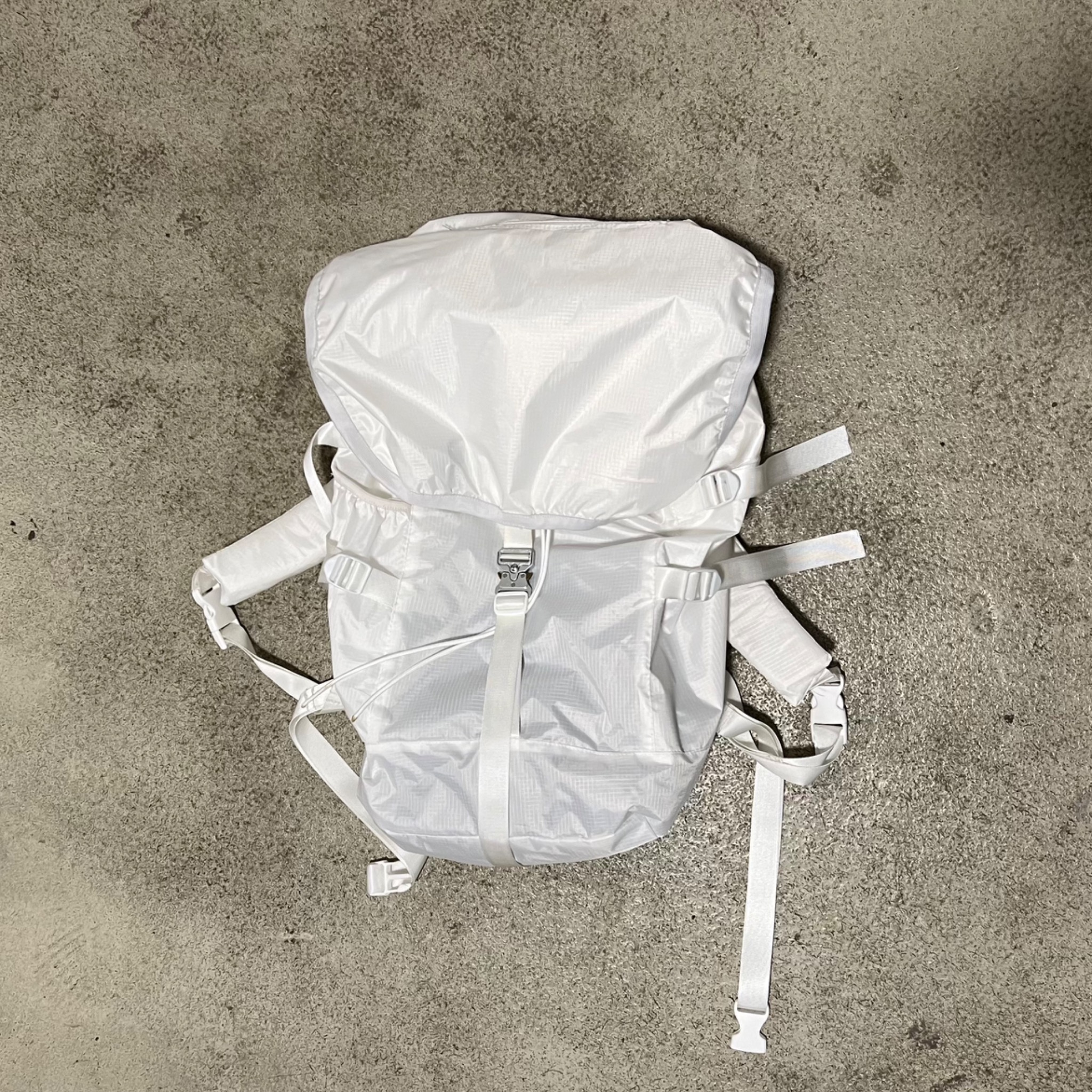 Biały, minimalistyczny plecak z lekkiego materiału, leżący na szarej, betonowej podłodze, widok z góry.