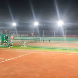 Oświetlony nocą kort tenisowy z ziemną nawierzchnią, siatką i wysokimi lampami oświetleniowymi, widoczne trybuny w tle.