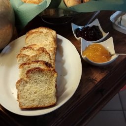 Wit - Al IWONA WITALSKA - Śniadanie na ciemnym drewnianym stole: kromki chałki na białym talerzu, miseczki z dżemem pomarańczowym i ciemnym owocowym, koszyk z bułeczkami pod szklanym kloszem, ceglana ściana w tle.