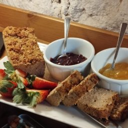 Wit - Al IWONA WITALSKA - Deser na białym talerzu: keks z kruszonką, kromki pieczywa, truskawki i dwa białe naczynia z dżemem wiśniowym i brzoskwiniowym, każda z łyżeczką.