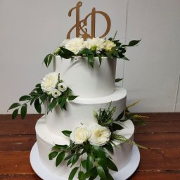 Wit - Al IWONA WITALSKA - Trzypiętrowy tort weselny z białym lukrem, ozdobiony białymi różami i zielonymi liśćmi, zwieńczony drewnianym topperem z inicjałami 'J&D'.
