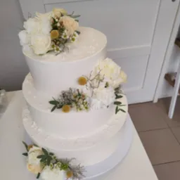 Dwupiętrowy tort weselny udekorowany białymi i kremowymi kwiatami oraz zielonymi listkami, umieszczony na białym stole, z widoczną podłogą z płytek w tle.