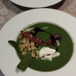 Porcja kremowej zielonej zupy na białym talerzu, udekorowana grzankami, bekonem, kleksem śmietany, sosem balsamicznym i zielonym strączkiem, na stole z koronkową serwetą.