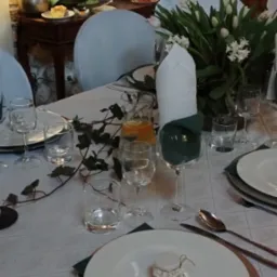 Uroczysty stół z białym obrusem w delikatny wzór, elegancką zastawą, bukietem białych tulipanów i hiacyntów, szklankami z wodą i plastrem pomarańczy oraz małym słoiczkiem z miodem na każdym talerzu.