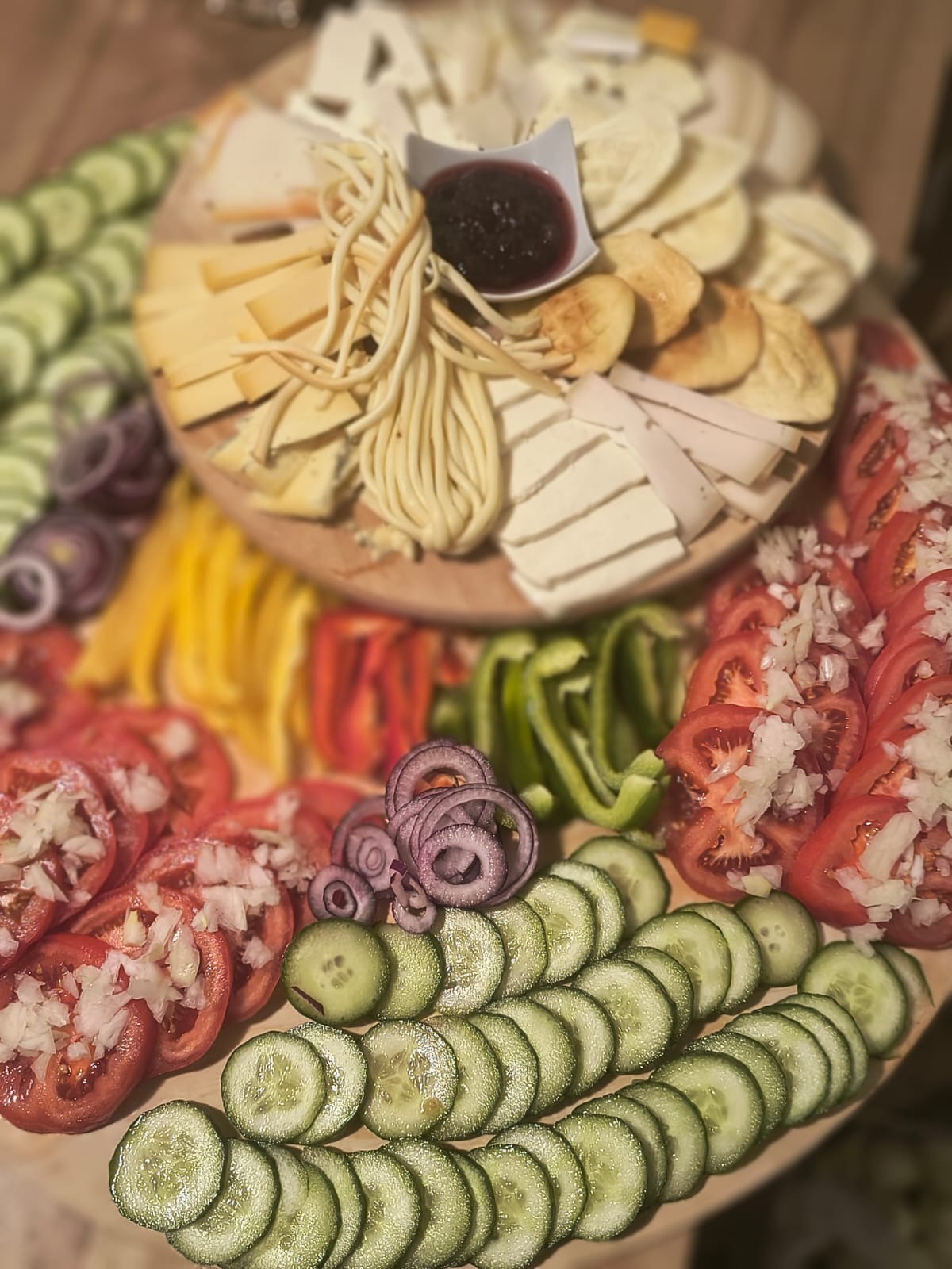 Dekoracyjna deska z serami, dżemem, pomidorami z cebulą, ogórkami, papryką i czerwoną cebulą, ułożona na drewnianej podstawie.