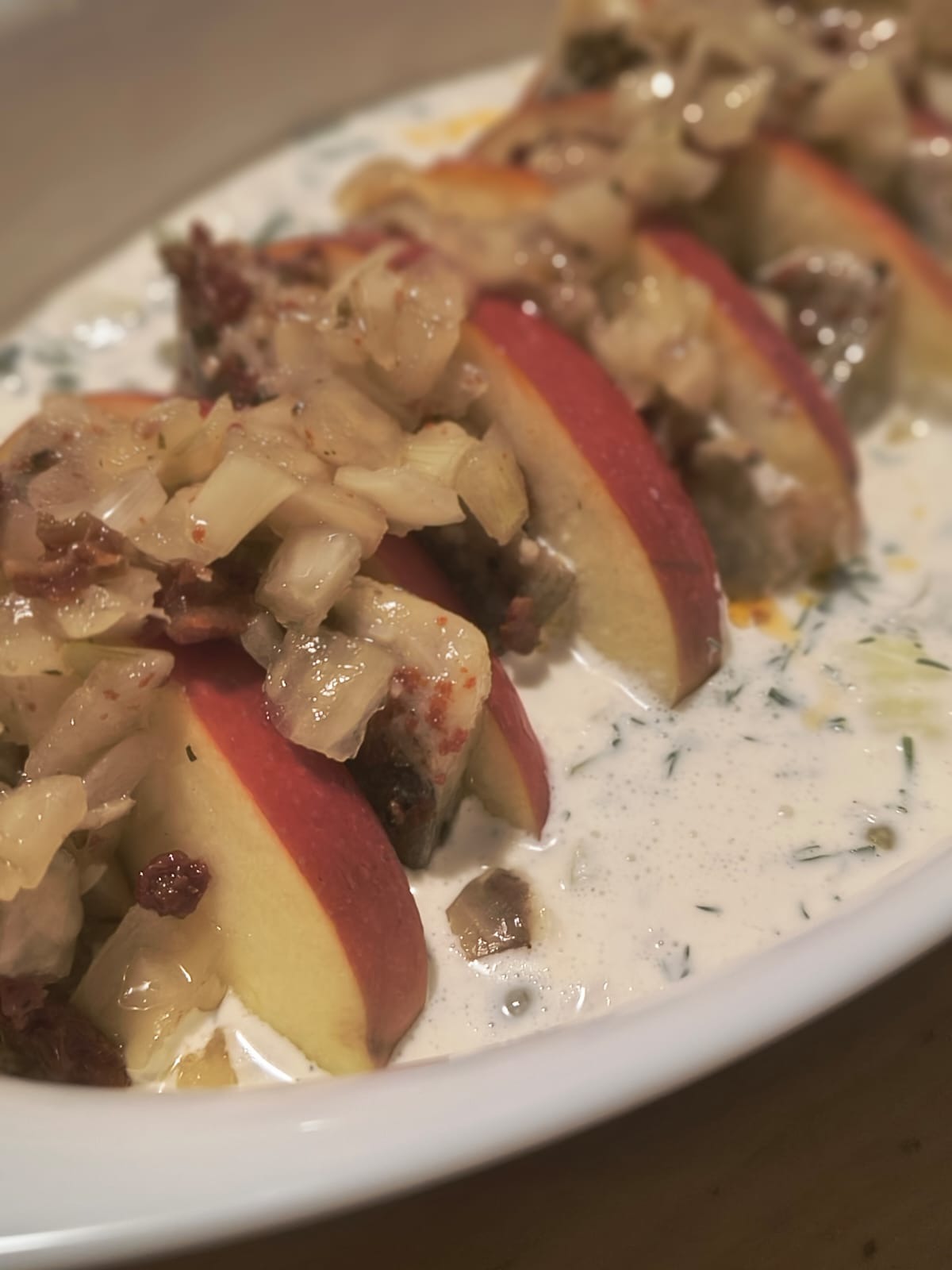 Jabłka w ćwiartkach ułożone w rzędzie, przykryte karmelizowaną cebulą i rodzynkami, zanurzone w kremowym sosie z zielonymi ziołami, podane na białym talerzu.